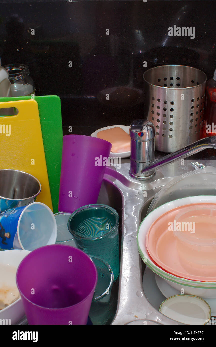 A collection of dirty dishes which includes plates, bowls, glasses, cutlery & chopping boards, stacked in a dirty kitchen sink with mixer tap. Stock Photo