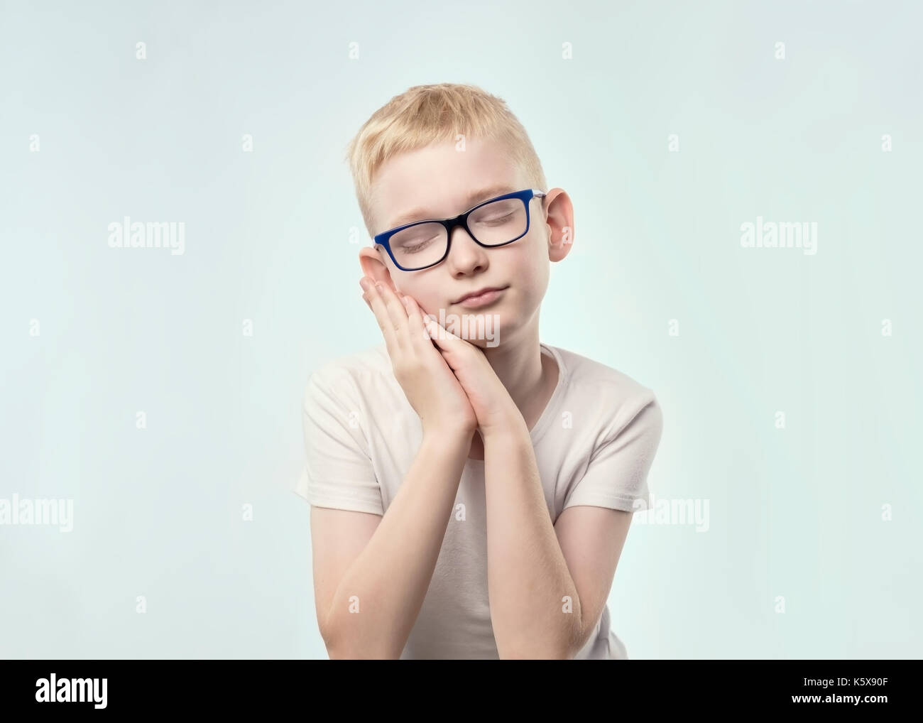 portrait of caucasian boy with blond hair. He is closing eyes and hold hands like pillow, wanting to sleep. Stock Photo