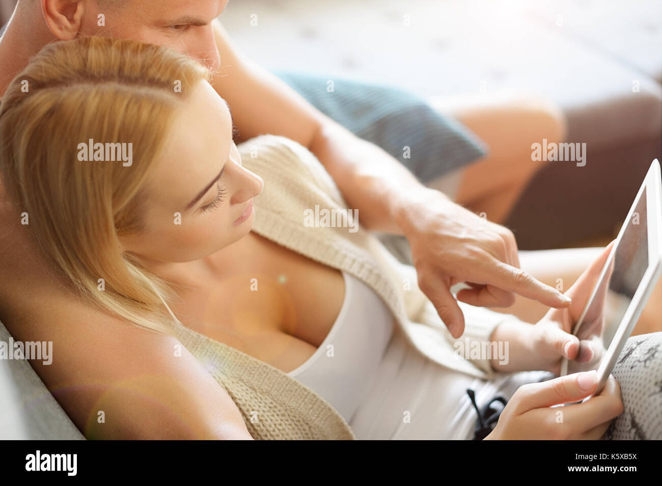 Young stylish couple cuddling on the sofa using digital tablet at their modern home. Woman holding tablet, browsing the web. Stock Photo