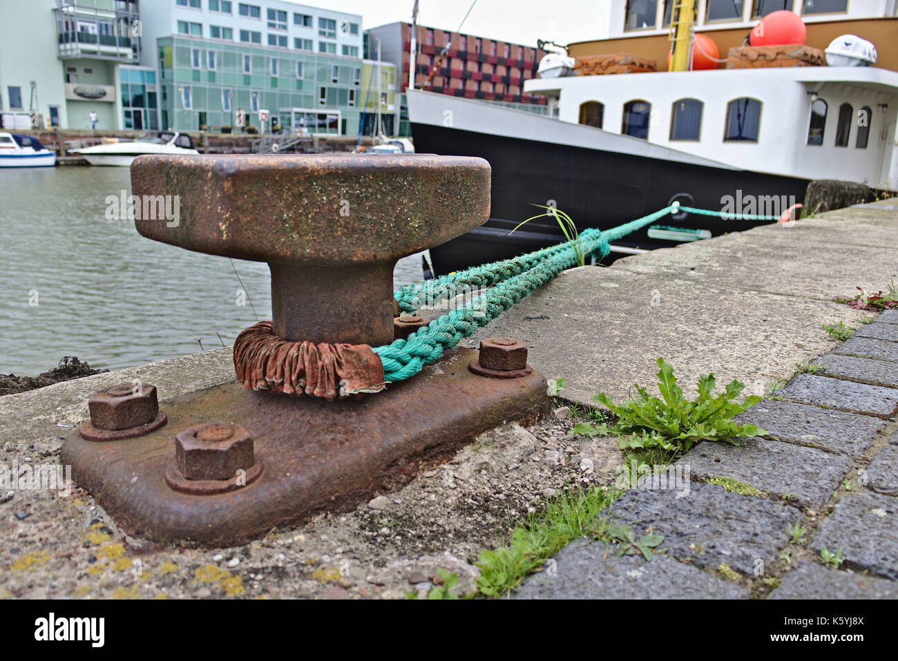 Rusty bollard with green mooring line leading to a ship alongside the pier and modern buildings