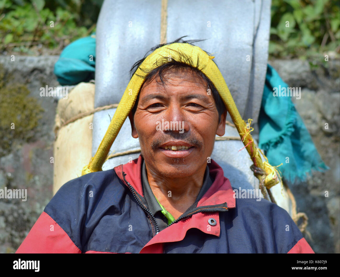 Nepali mountain porter (sherpa) with a heavy load takes a short break on a stone bench and smiles for the camera. Stock Photo