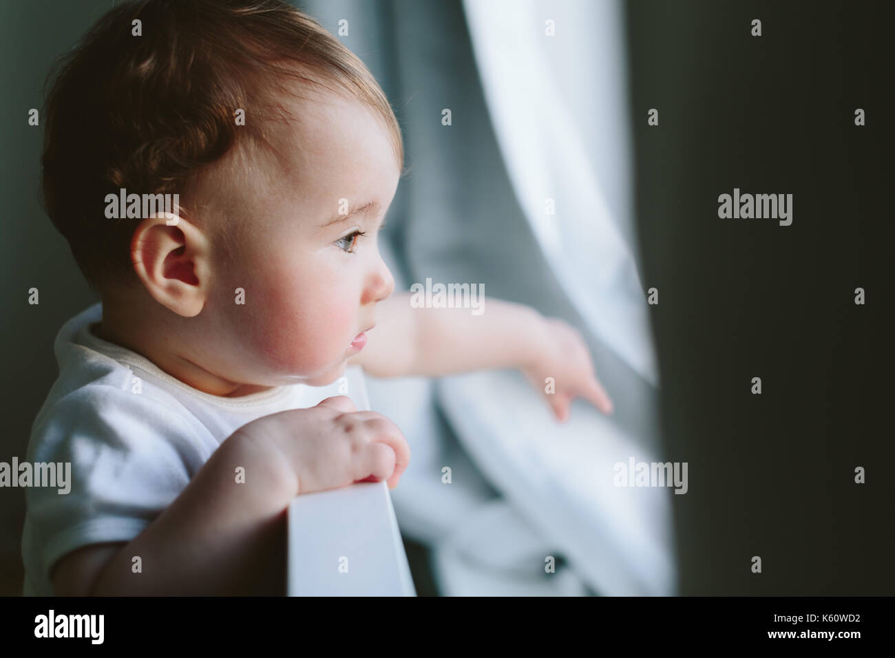 Little baby holds onto the bed and looks somewhere. Stock Photo