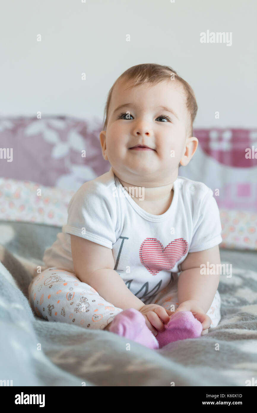 Vertical shot. Little baby smiling sits on bed at home. Stock Photo