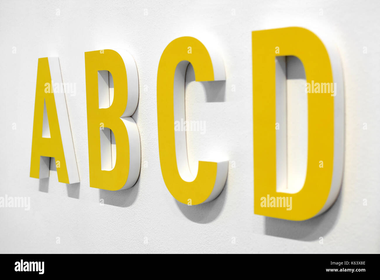 ABCD yellow alphabet letters mounted on a white wall with shadow viewed at an oblique angle with D in the foreground Stock Photo