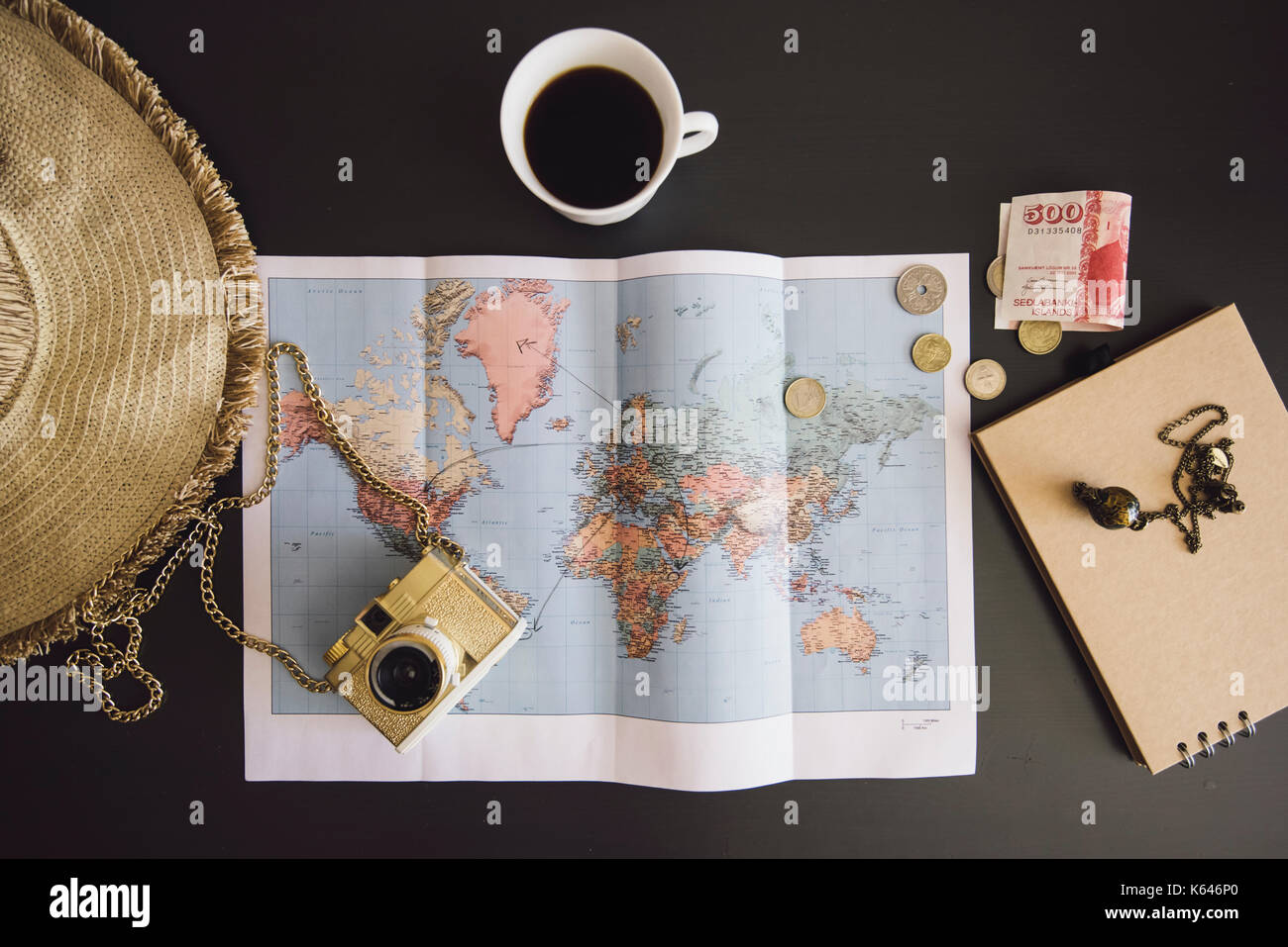 Planning the trip. World map with the hat, film camera, some money, notebook from recycled paper and freshly brewed coffee cup on the dark table Stock Photo