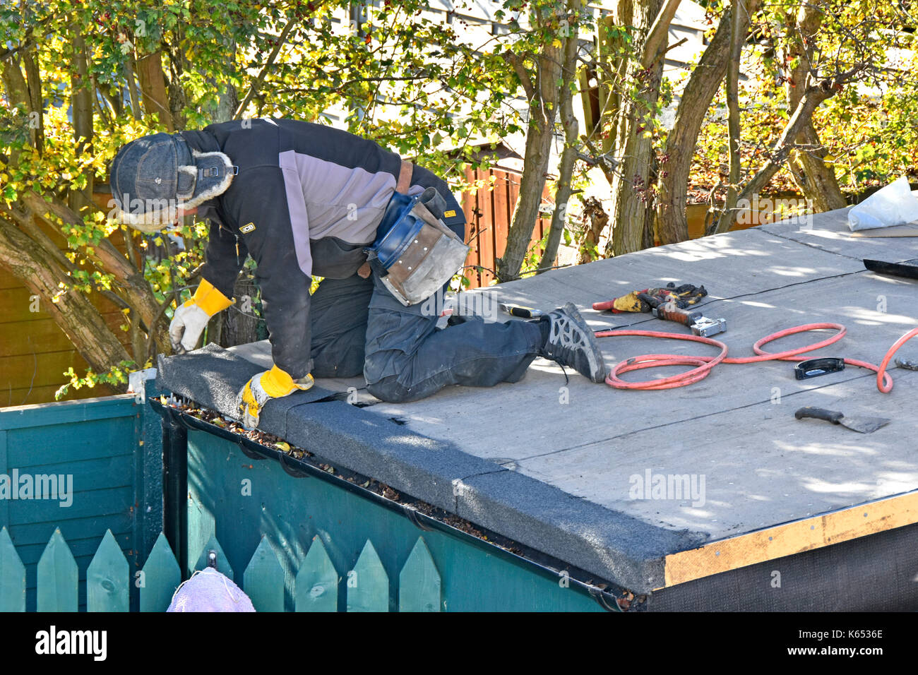 Roofer working on re-covering leaking flat garden shed roof pressing down on heated bitumen adhesive on new roofing felt Stock Photo