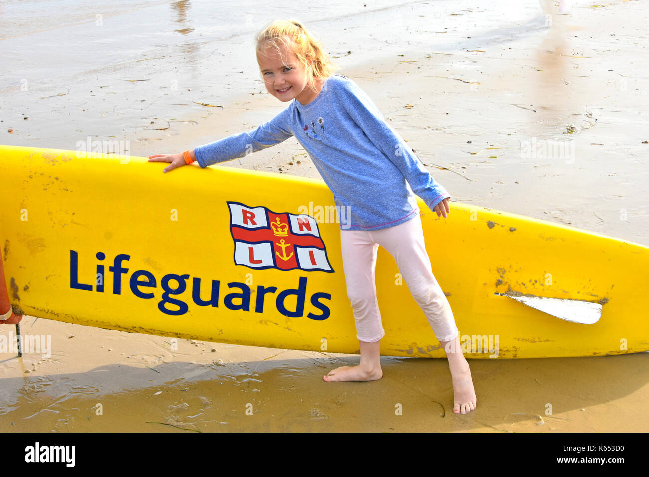 Six year old girl posing at seaside beside a RNLI Lifeguards surfboard awareness idea of sea bathing hazards chilly summer holiday to Dorset beach UK Stock Photo