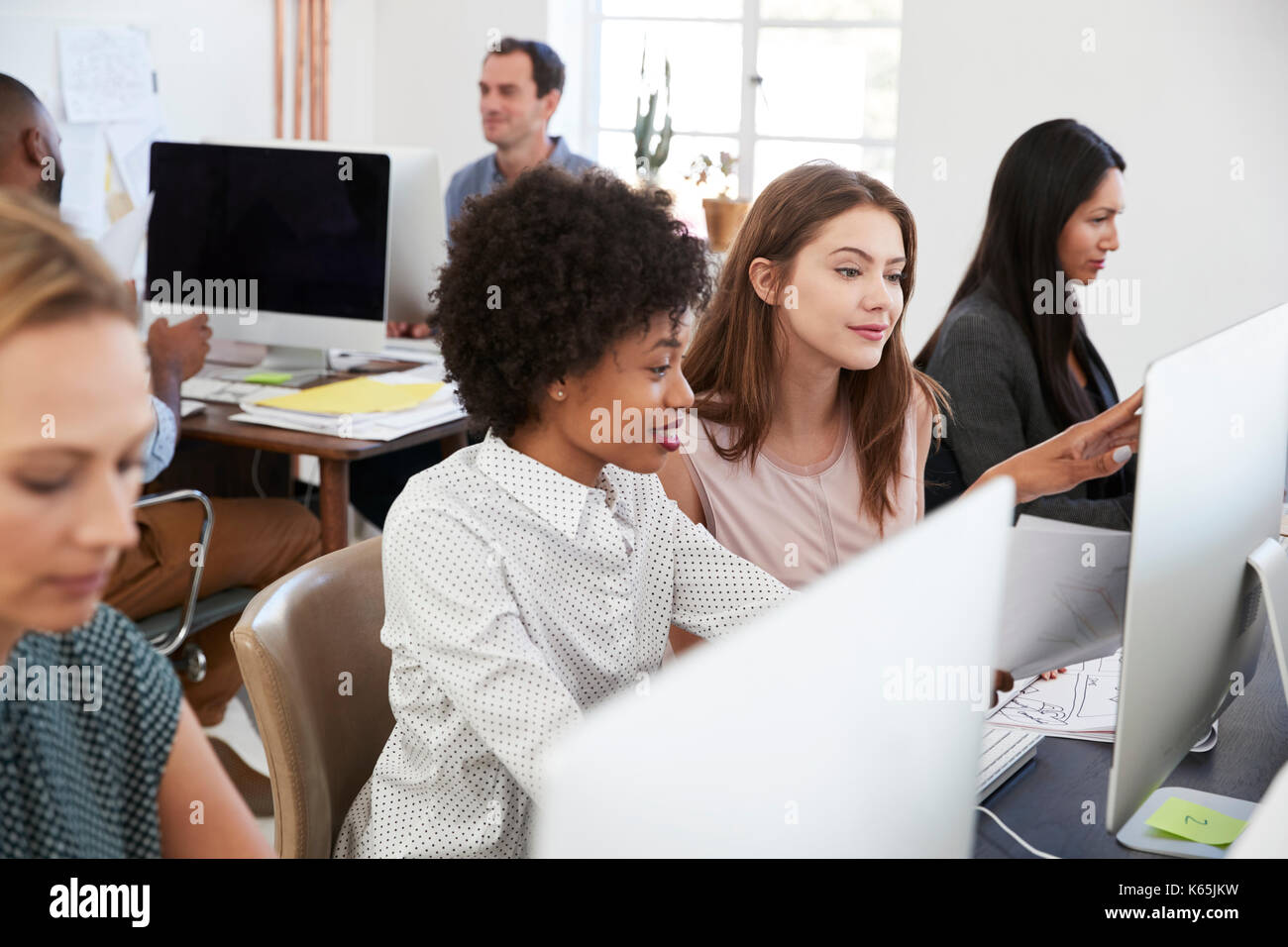Two women sat at a computer discuss work in open plan office Stock Photo