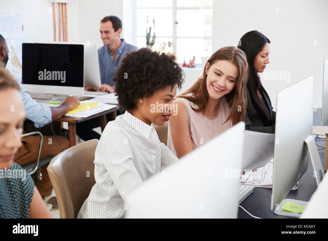 Two happy women discuss work at computer in open plan office Stock Photo