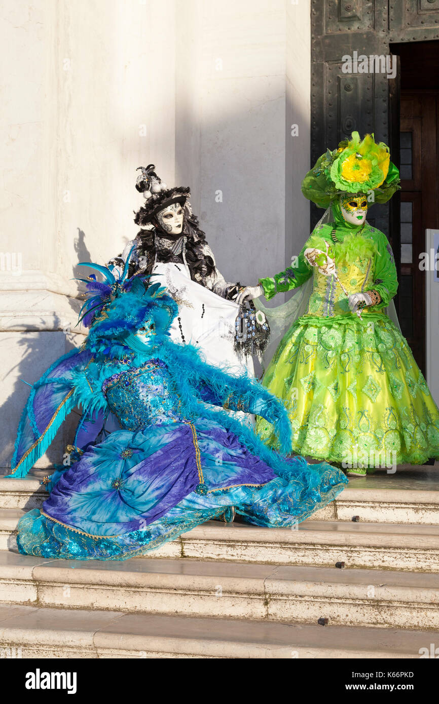 Venice Carnival 2017, Italy. Three brightly colored women in classical costume and masks pose on the steps of San Giorgio Maggiore in late evening lig Stock Photo