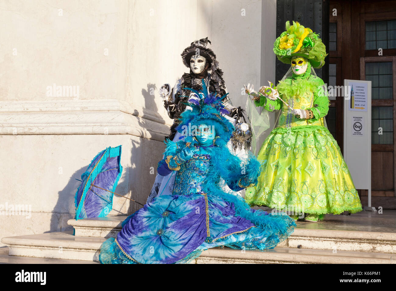 Venice Carnival 2017, Italy. Three colorful women in classical costume and masks pose on the steps of San Giorgio Maggiore in late evening light with  Stock Photo