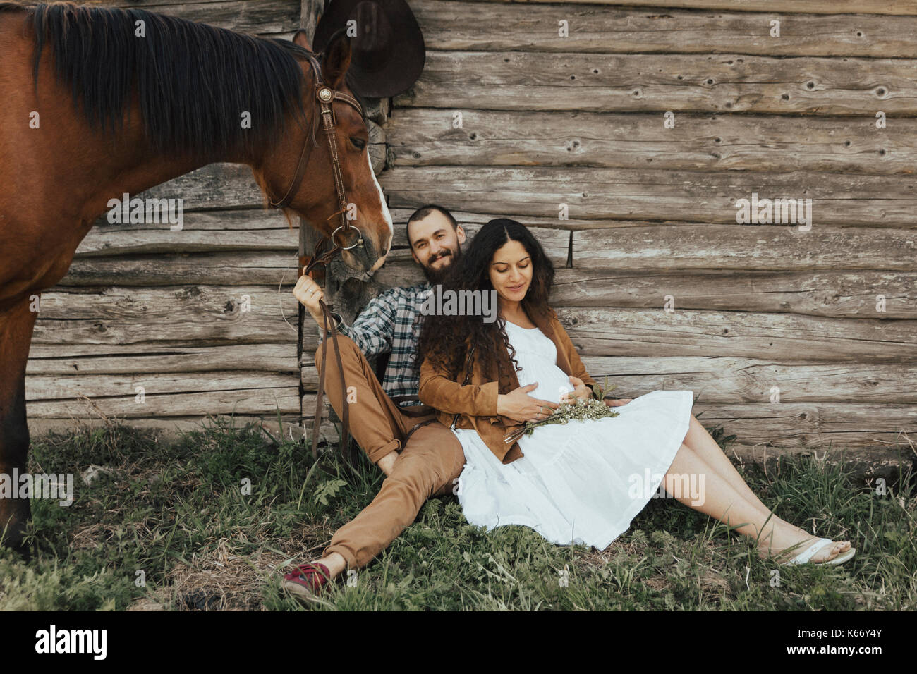Couple sitting in grass near barn with horse Stock Photo