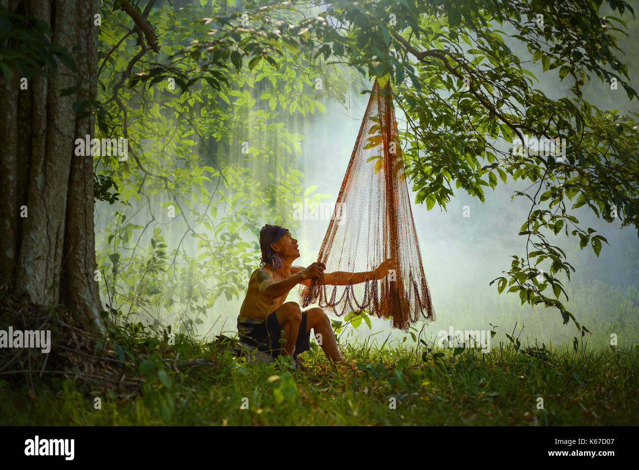 Fisherman sitting in forest inspecting fishing net, Thailand Stock Photo