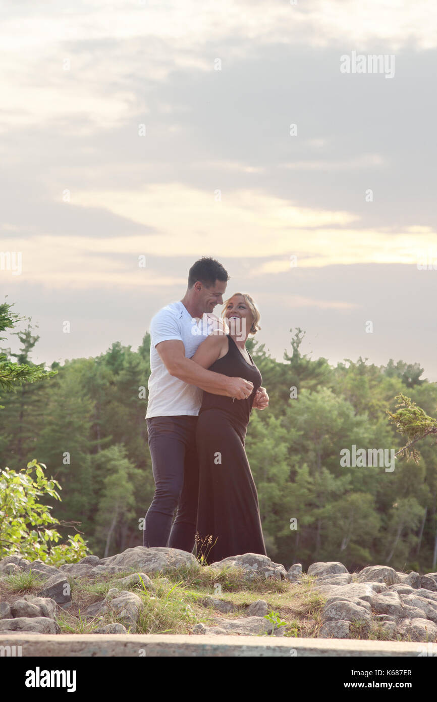 a beautiful blond and handsome man stand on a rock outside, laughing and in love, with the sun set behind Stock Photo