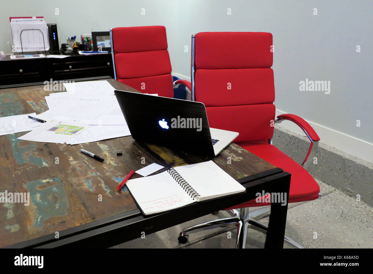 Two red office chairs sitting at a table with a mac book laptop sitting open beside a stack of papers. Stock Photo
