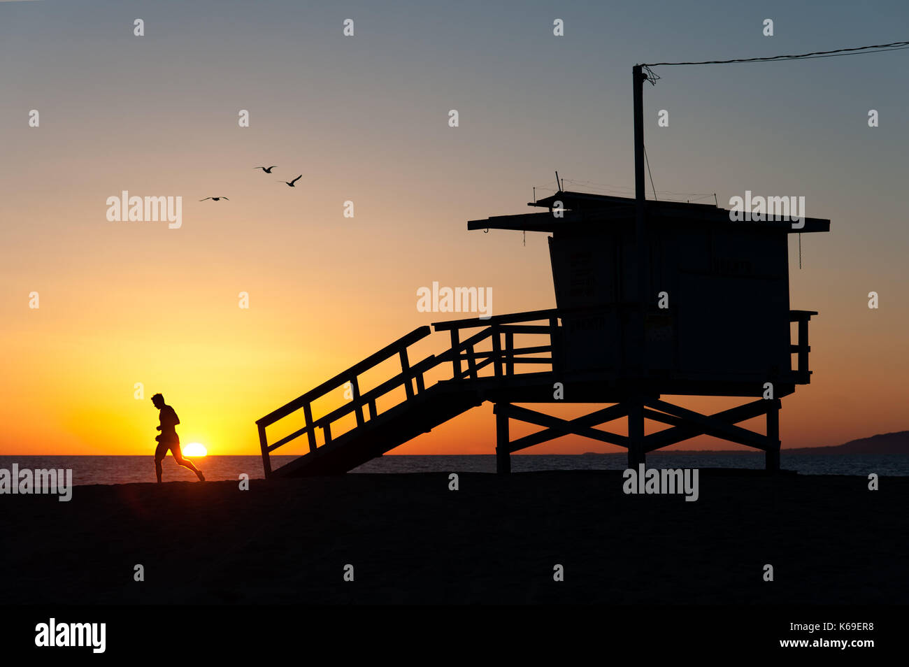 Silhouettes of a sportive runner working out on the sand and the iconic lifeguard tower with flying birds at sunset in Venice Beach, CA. Stock Photo