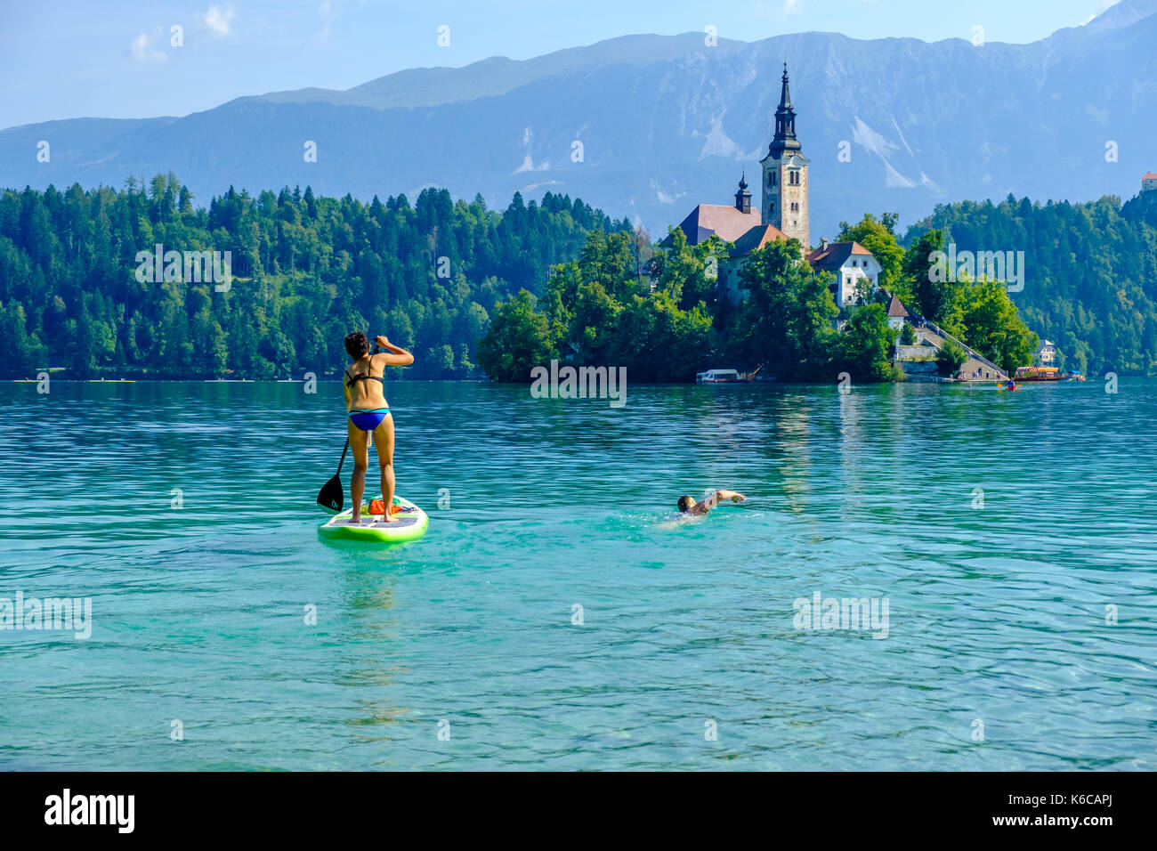 A young woman is stand up paddling on Lake Bled, Blejsko jezero, another woman is swimming next to her towards Bled Island, Blejski otok Stock Photo