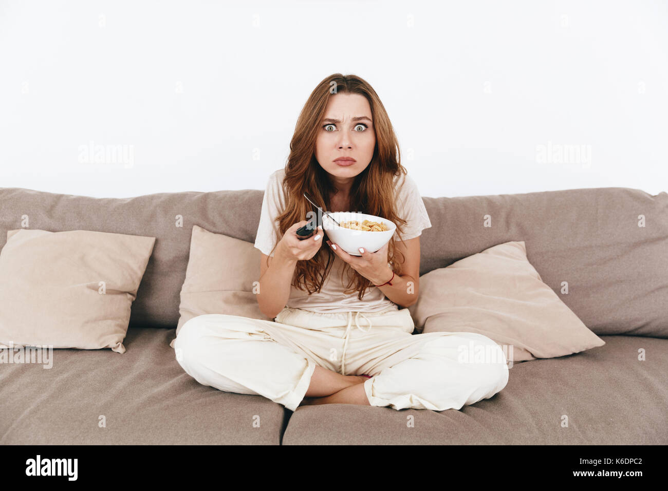 Picture of young concentrated amazing pretty lady sitting on sofa indoors. Looking camera eating corn flakes holding remote control watch TV. Stock Photo