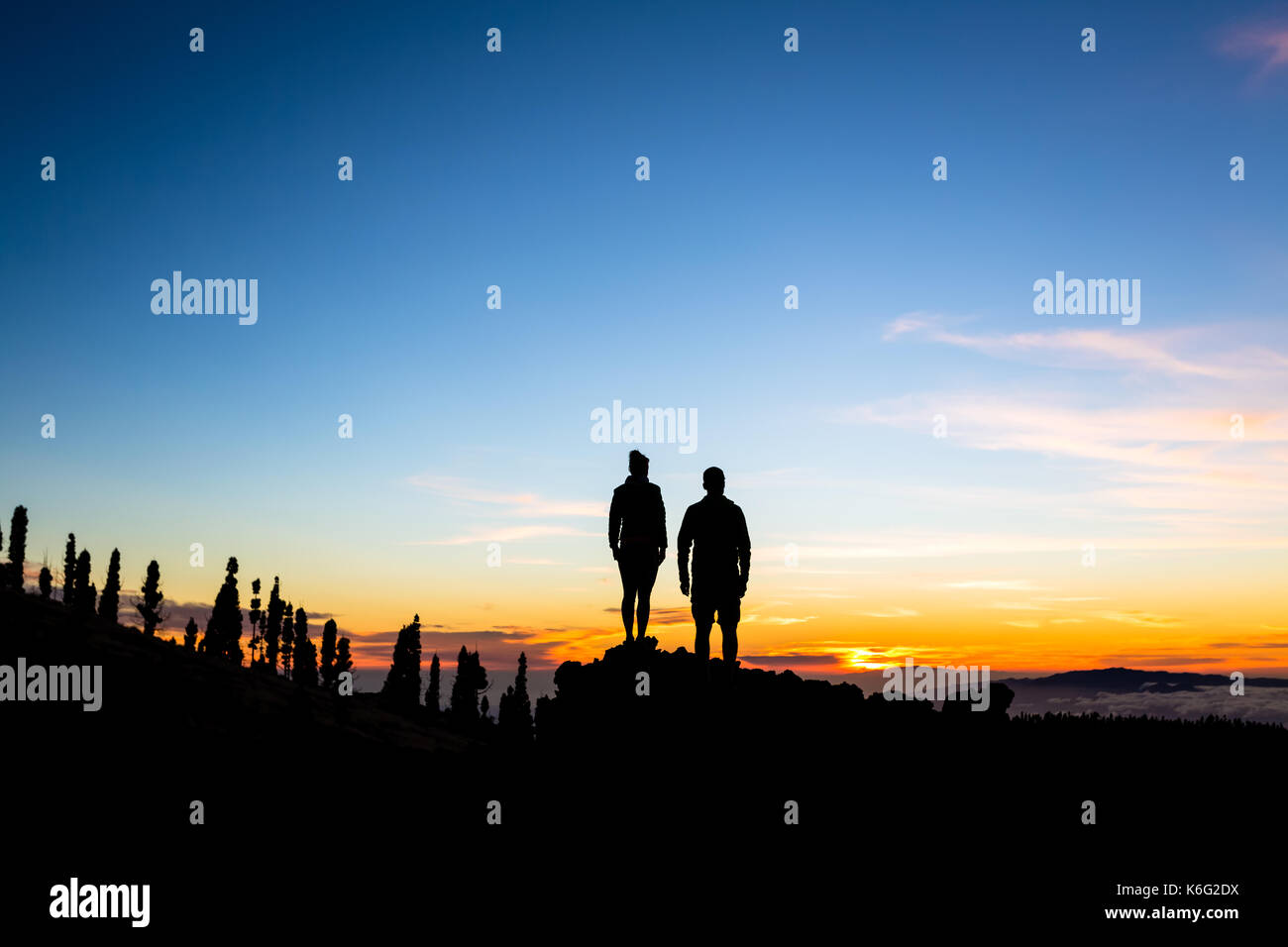 Teamwork couple climbing and reaching mountain peak. Silhouette of climbers team over mountains sunset. Man and woman hikers looking at inspirational  Stock Photo