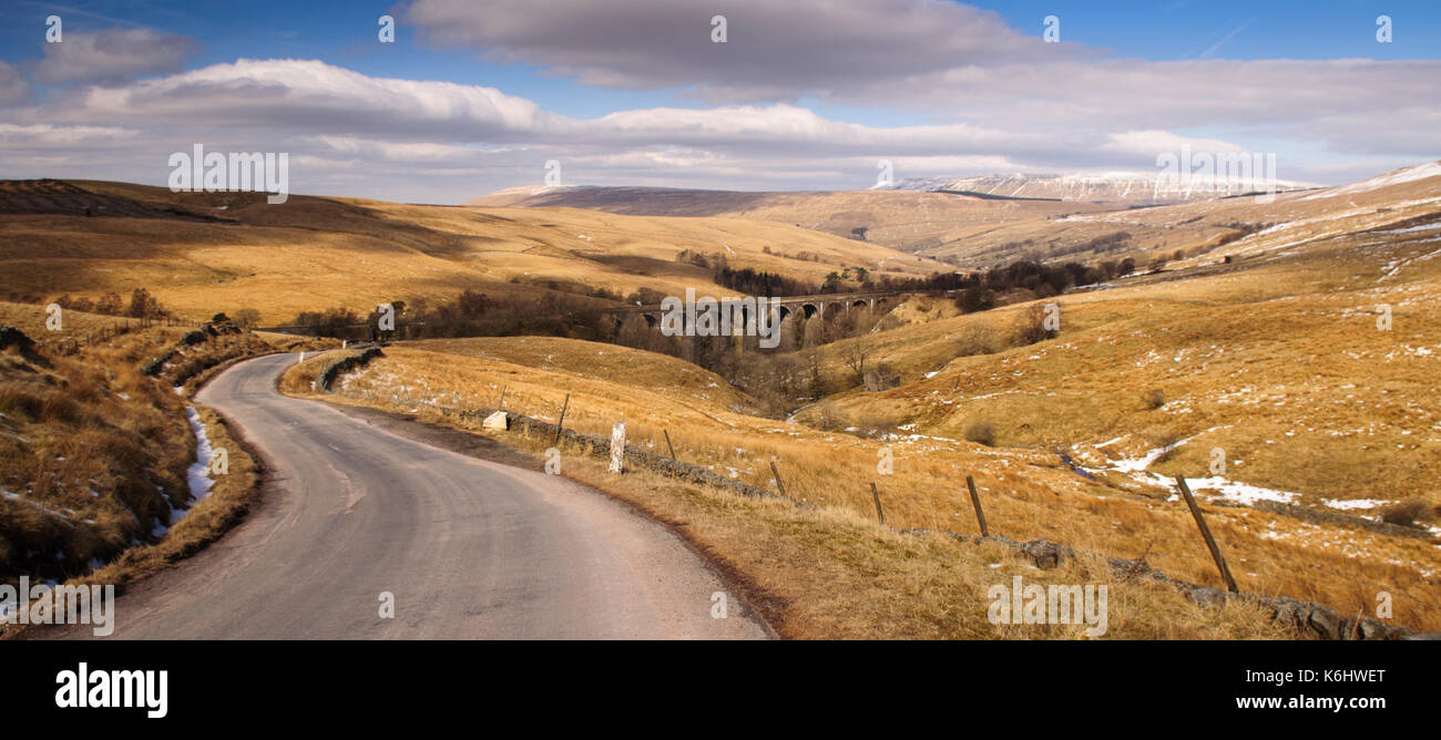 A single track country lane climbs from Dentdale above the Dent Head Viaduct on the Settle-Carlisle Railway Line in the rolling moorland landscape of  Stock Photo