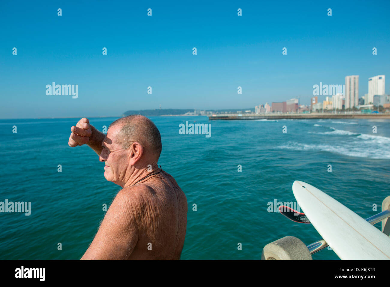 Senior man with surfboard watching for waves on pier near promenade on Golden Mile, Durban, South Africa Stock Photo
