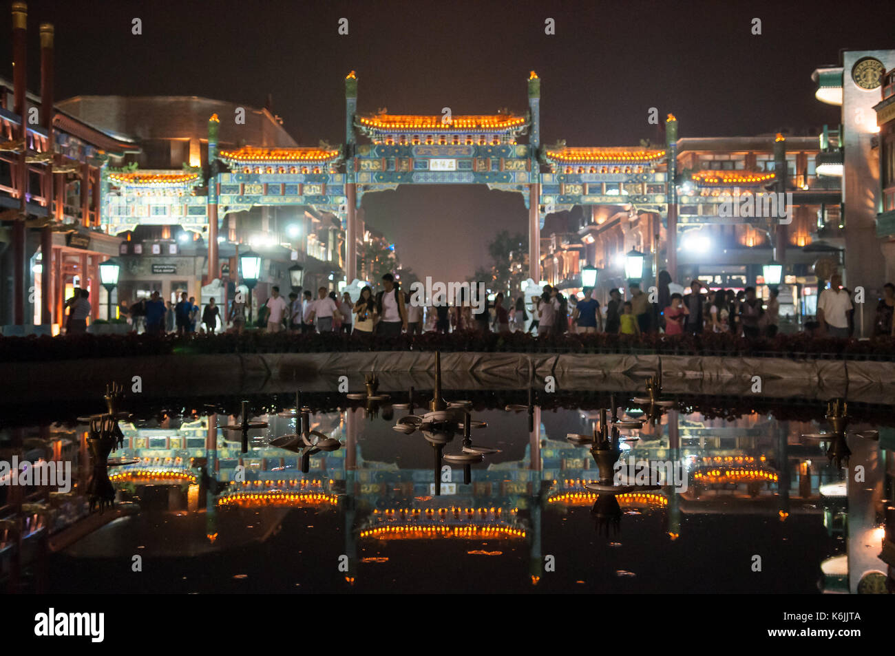 Crowds of pedestrians walk through the traditional Chinese Paifang gateway at the entrance to Qianmen Street, reflected in the waters of a fountain at Stock Photo