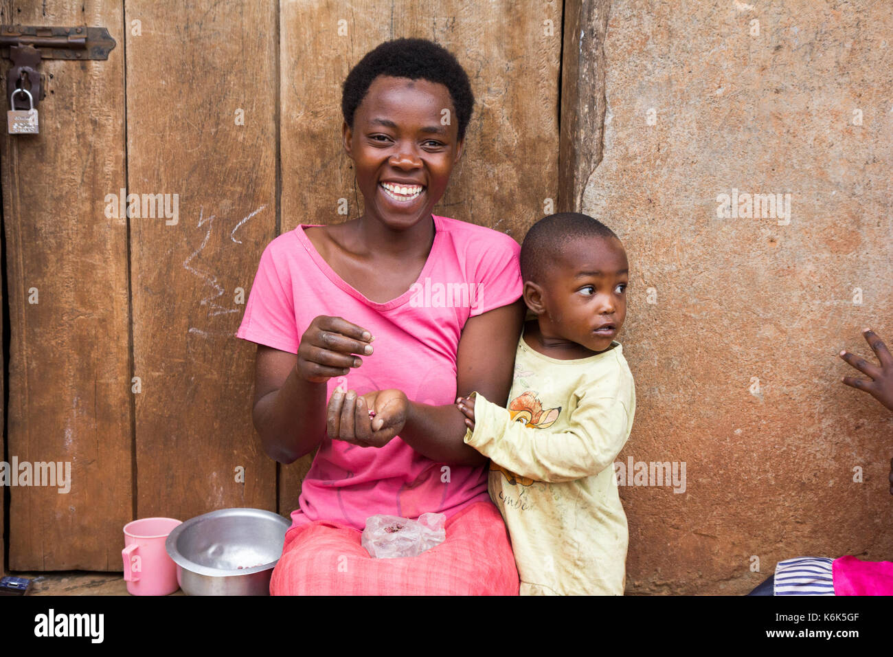 Lugazi, Uganda. 09 June 2017. A laughing African mother with a little child boy. The woman is sitting on door threshold sorting grains. Stock Photo