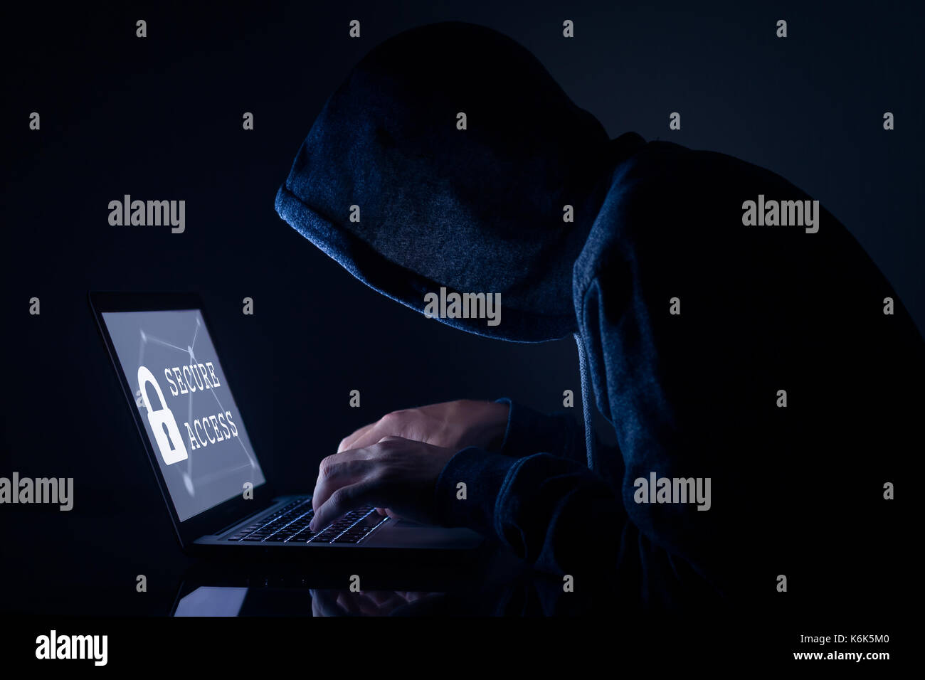 Hooded hacker in dark room performing a cyber attack to a secure access to steal data on internet, laptop computer screen Stock Photo