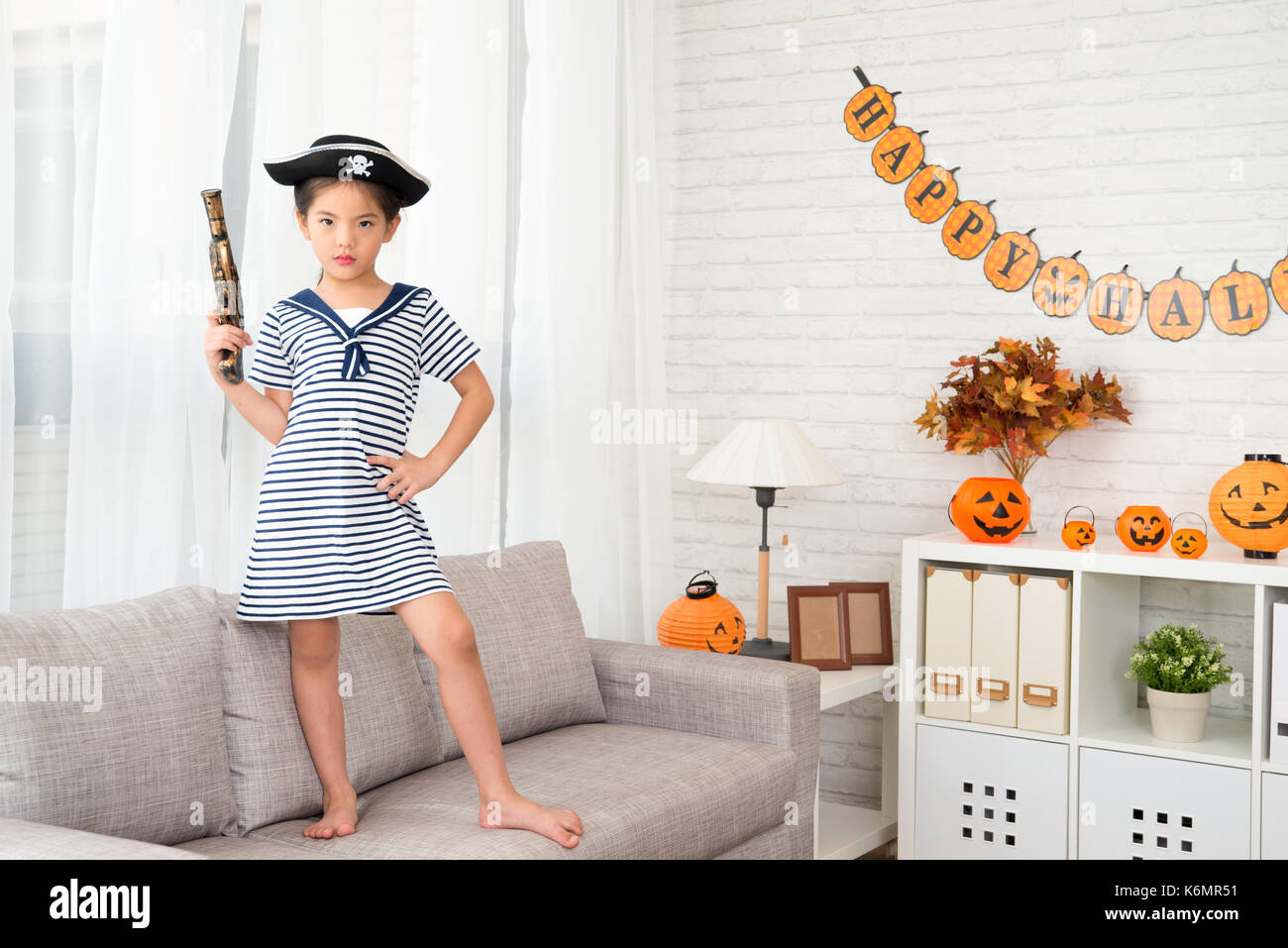 serious confidence naughty captain girl holding a pirate toy pistol have fun for Halloween holiday standing on sofa at home Stock Photo