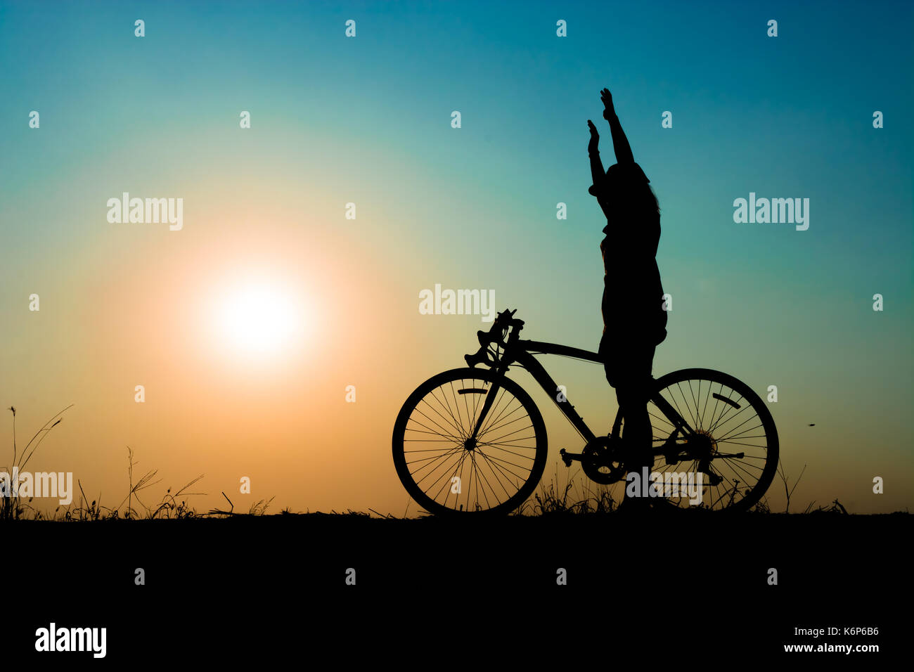 silhouette of a girl and her bike on meadow at the sunset time. Standing and hand up while riding. Have a beauty blue sky and white - pink of a sun. Stock Photo