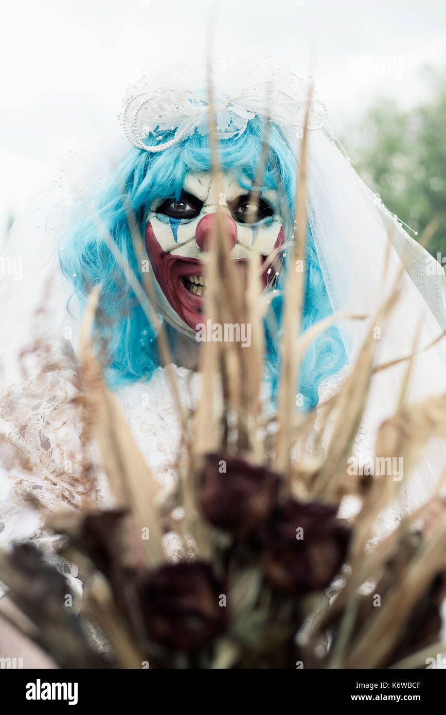 closeup of a scary evil clown wearing a dirty and ragged bride dress holding a bridal bouquet with wilt flowers in front of the observer Stock Photo