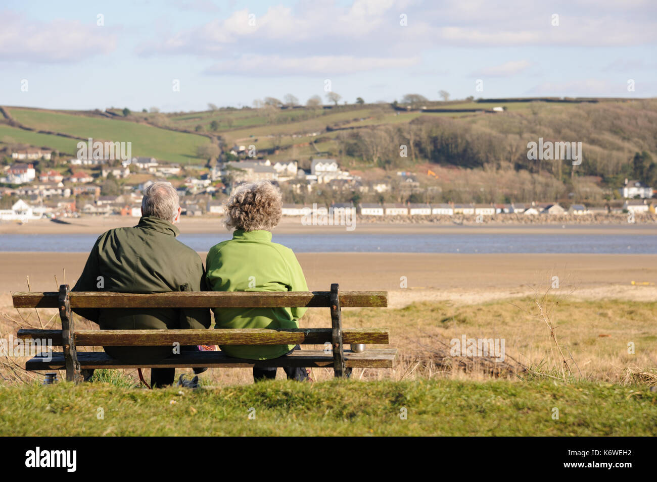 A couple sit on a bench facing the small village of Ferryside on the oposite shore in the distance and overlooking the river Towy, Carmarthenshire. Wa Stock Photo