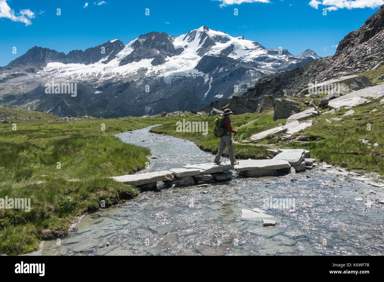 Hiker crosses mountain stream, alpine meadow Valle delle Meyes, 2700 m a. s. l., roundwader trail from Pont, panorama of the Stock Photo