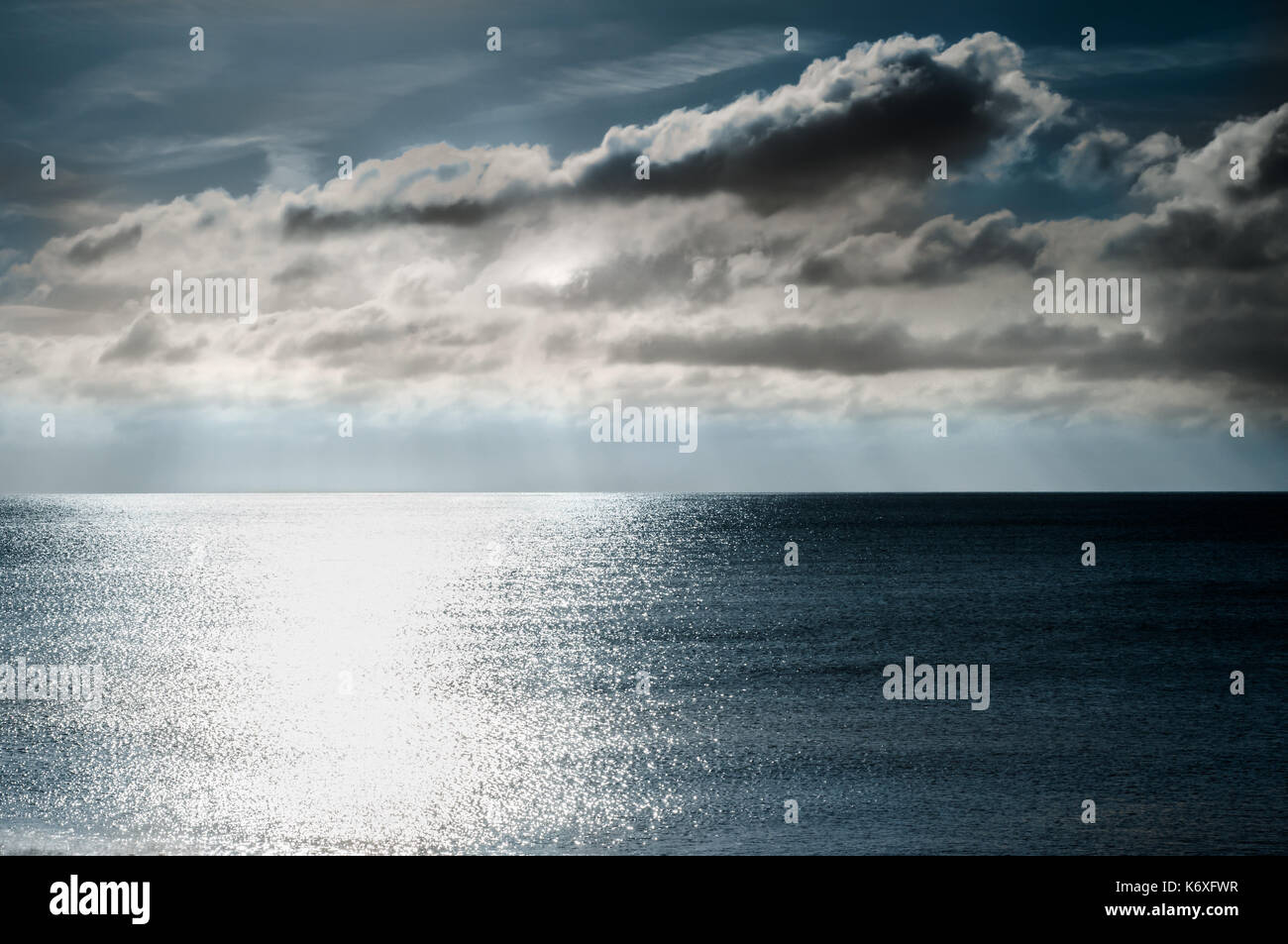 Seascape at dusk, with grey clouds in a dark moody sky and sunlight rays shining and scattering on dark blue water. Stock Photo