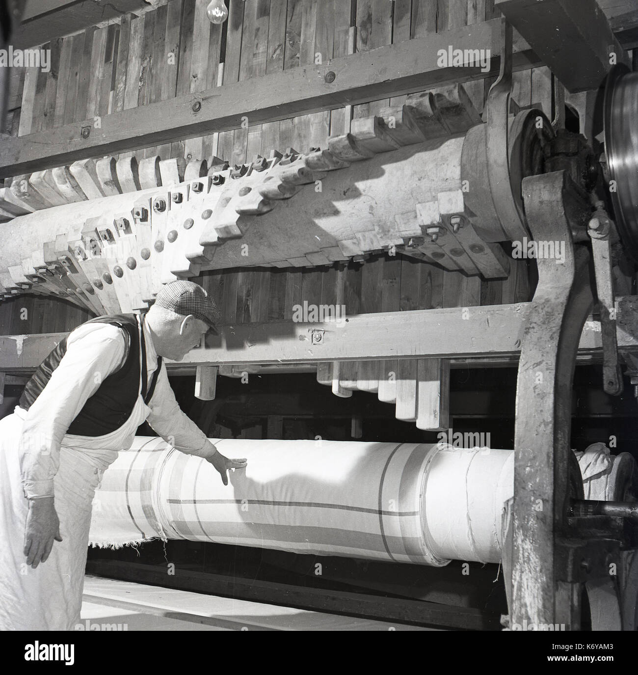 1950s, historical, a male textile worker checking the quality of the finished linen cloth tightly wrapped on giant wooden rollers, Belfast, Northern Ireland. Irish linen is famous around the world, as the weather and land conditions help produce the flax plant (weed) that the cloth is made from. Stock Photo
