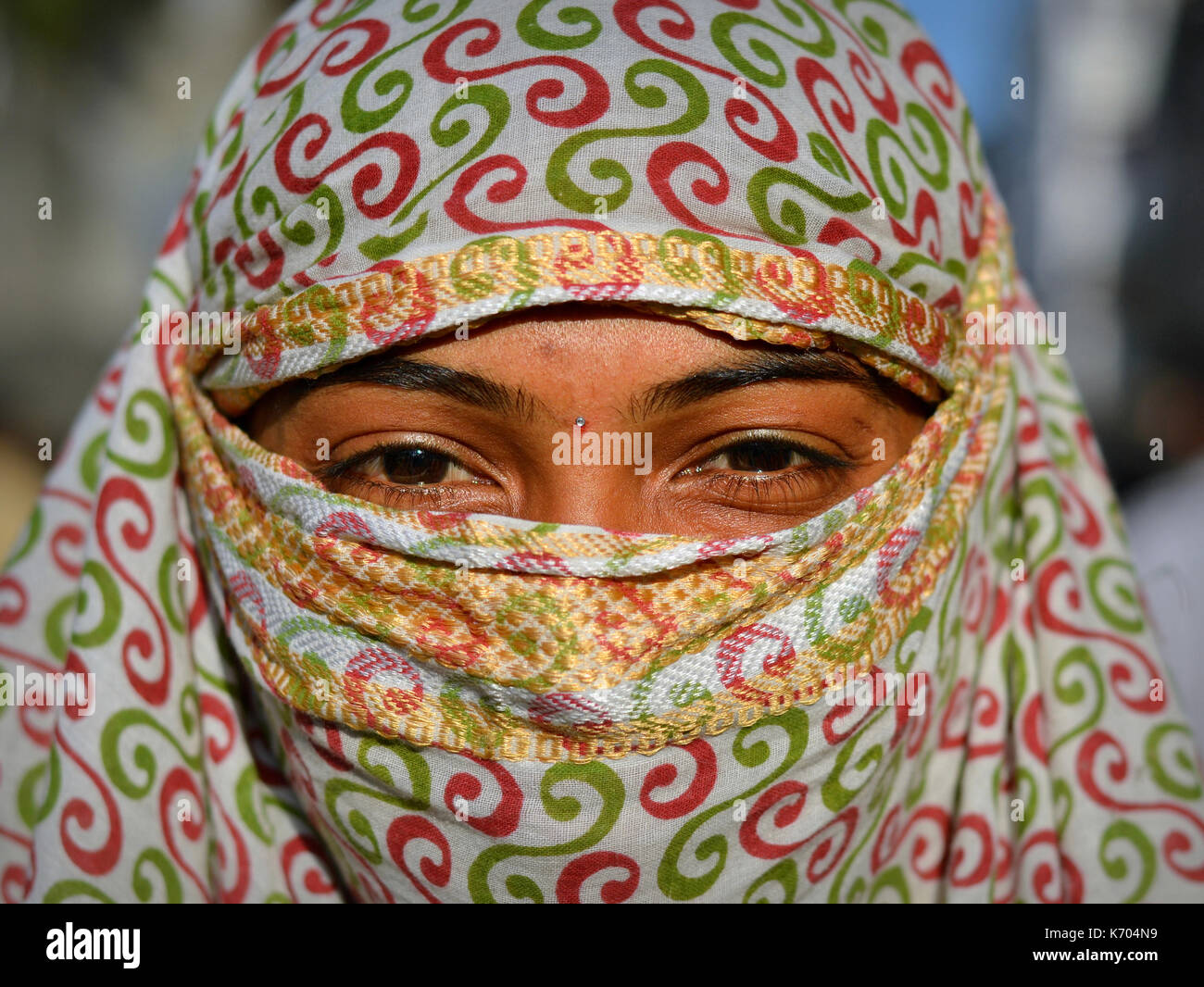 Young Indian woman with beautiful eyes covers her hair and face with a trendy secular, red-and-green headscarf and poses for the camera. Stock Photo