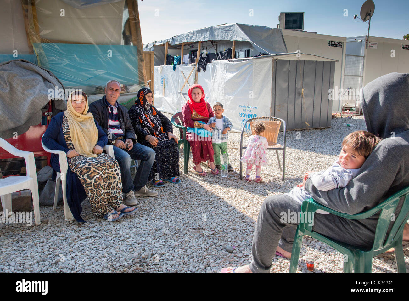 A group of Syrian Refugees, ranging from grandmotherly age to toddlers gather sociably on plastic chairs, between the Isobox houses at Ritsona Camp, Stock Photo
