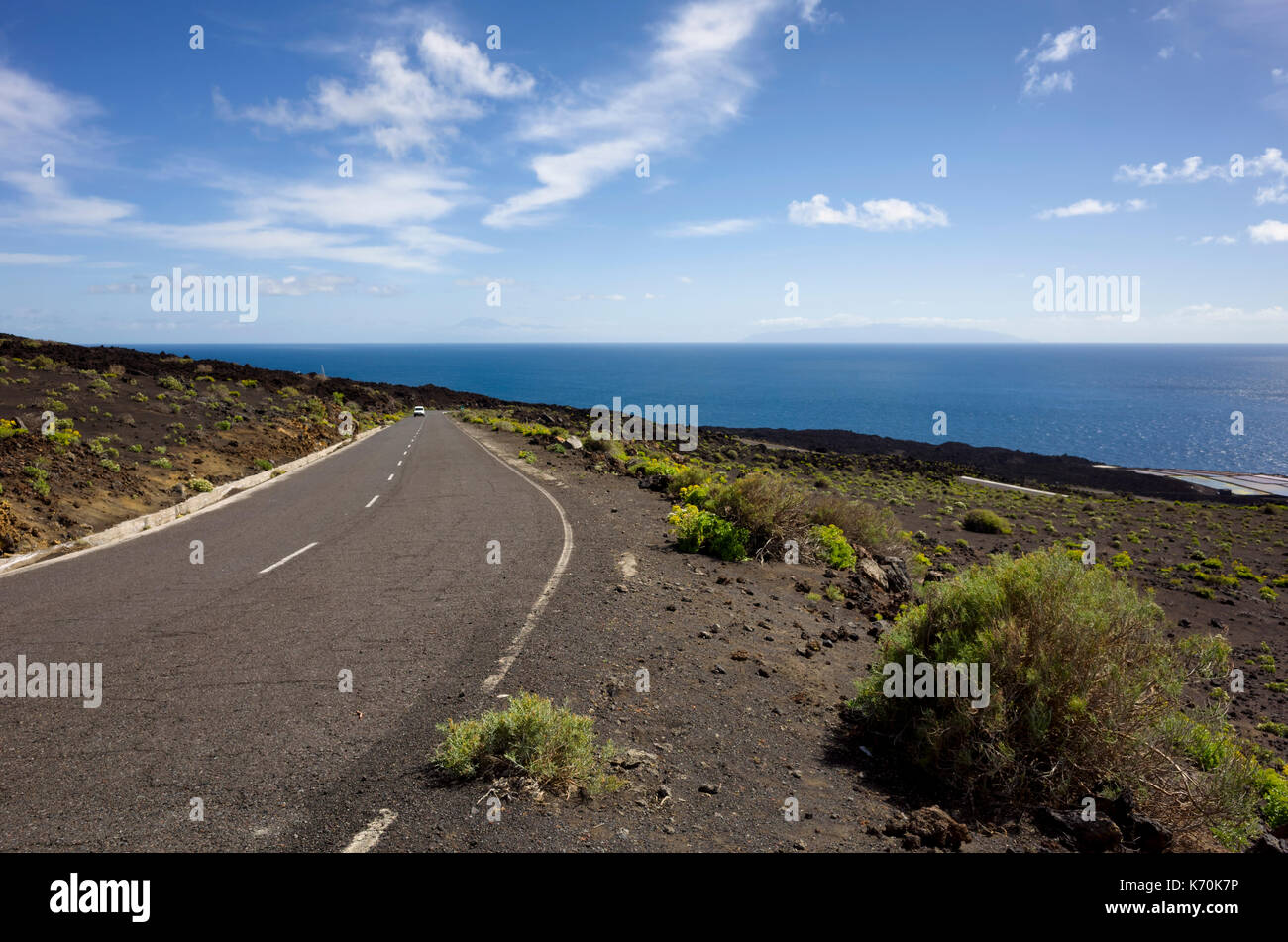 LP-130 highway, Salinas de Fuencaliente, La Palma.  A view of the coastal road, close to sea level, where it comes to the tourist attractions of the two light houses and sea salt pans. A little vegetation grows along the road side.  Lava rocks form the coastline and a beautiful deep blue sea can be seen to the horizon against a blue sky with light fluffy clouds.  The neighbouring island is just about visible on the horizon. Stock Photo