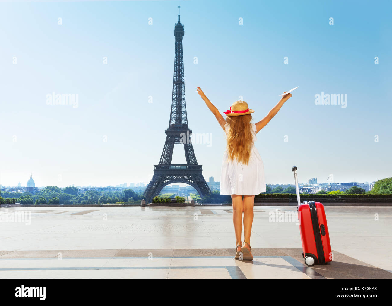 Back view portrait of little girl, wearing straw hat and white dress, looking at the Eiffel Tower with her hands lifted up Stock Photo