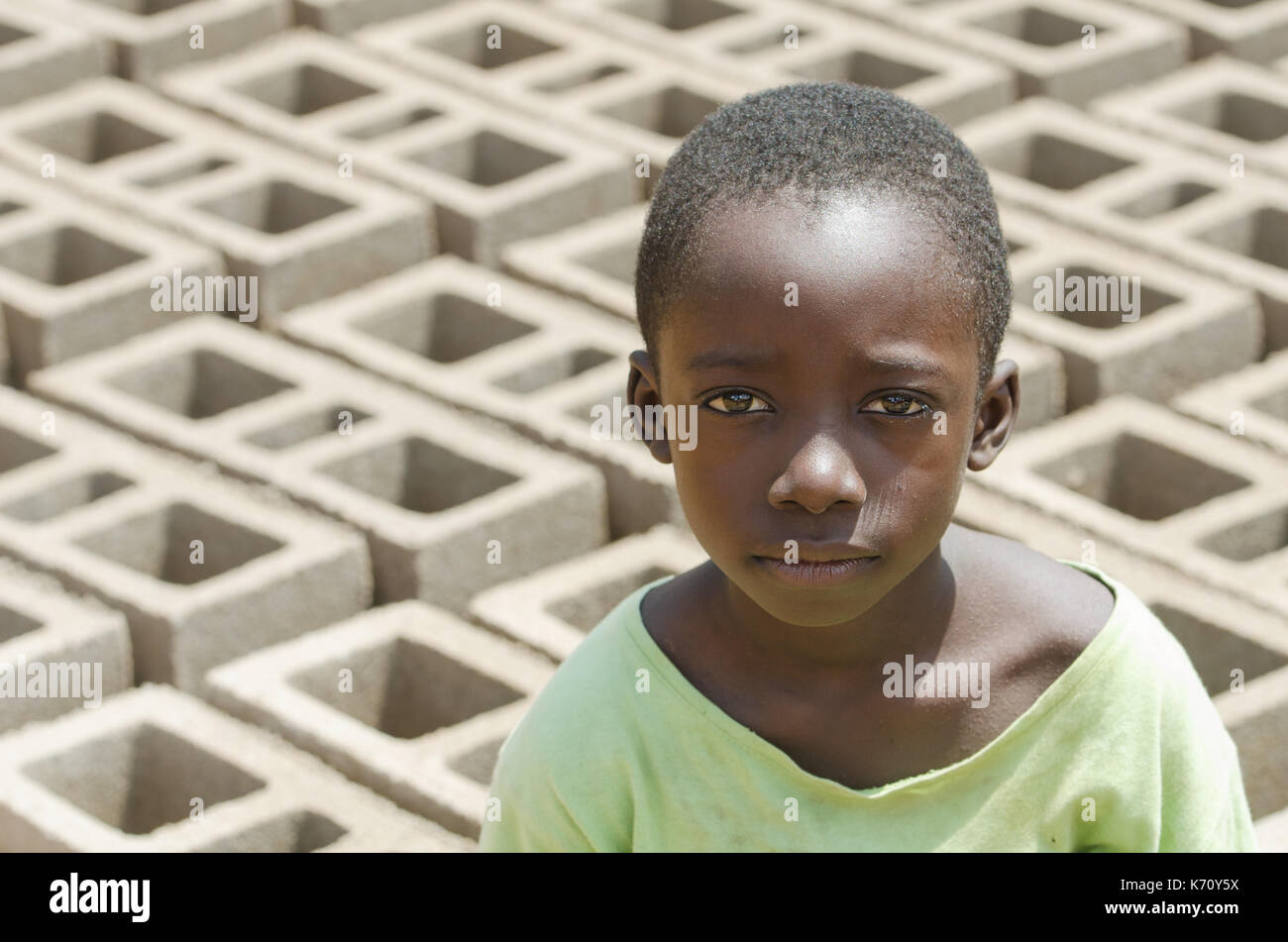 Black child sitting outdoors as a child labor concept - Isolated in front of a lot of bricks to build Stock Photo
