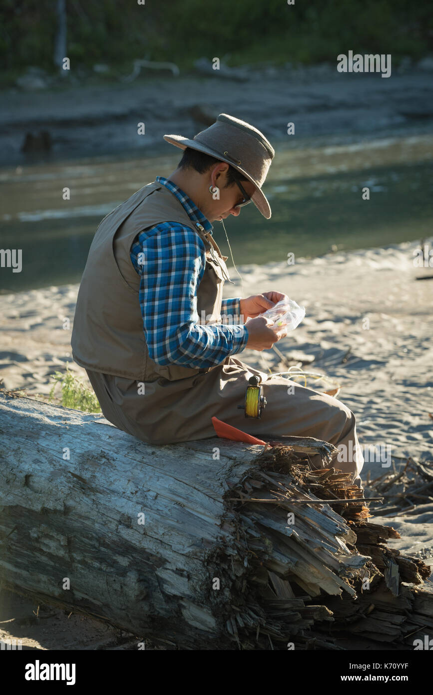Side view of man looking at fishing bait while sitting on fallen tree trunk during sunny day Stock Photo