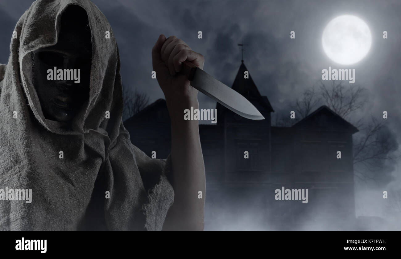 Hooded man with big knife . Abandoned wooden house in the moonlit night . Halloween background. Stock Photo