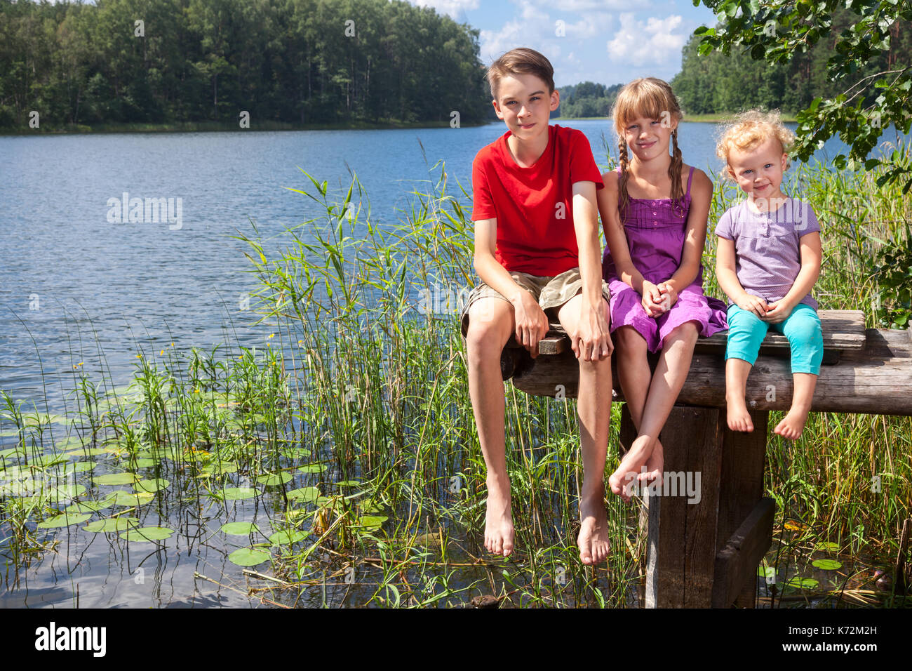 Three children of different age - teenager boy, elementary age girl and toddler girl sitting on a wooden pier by a forest lake looking at camera smili Stock Photo