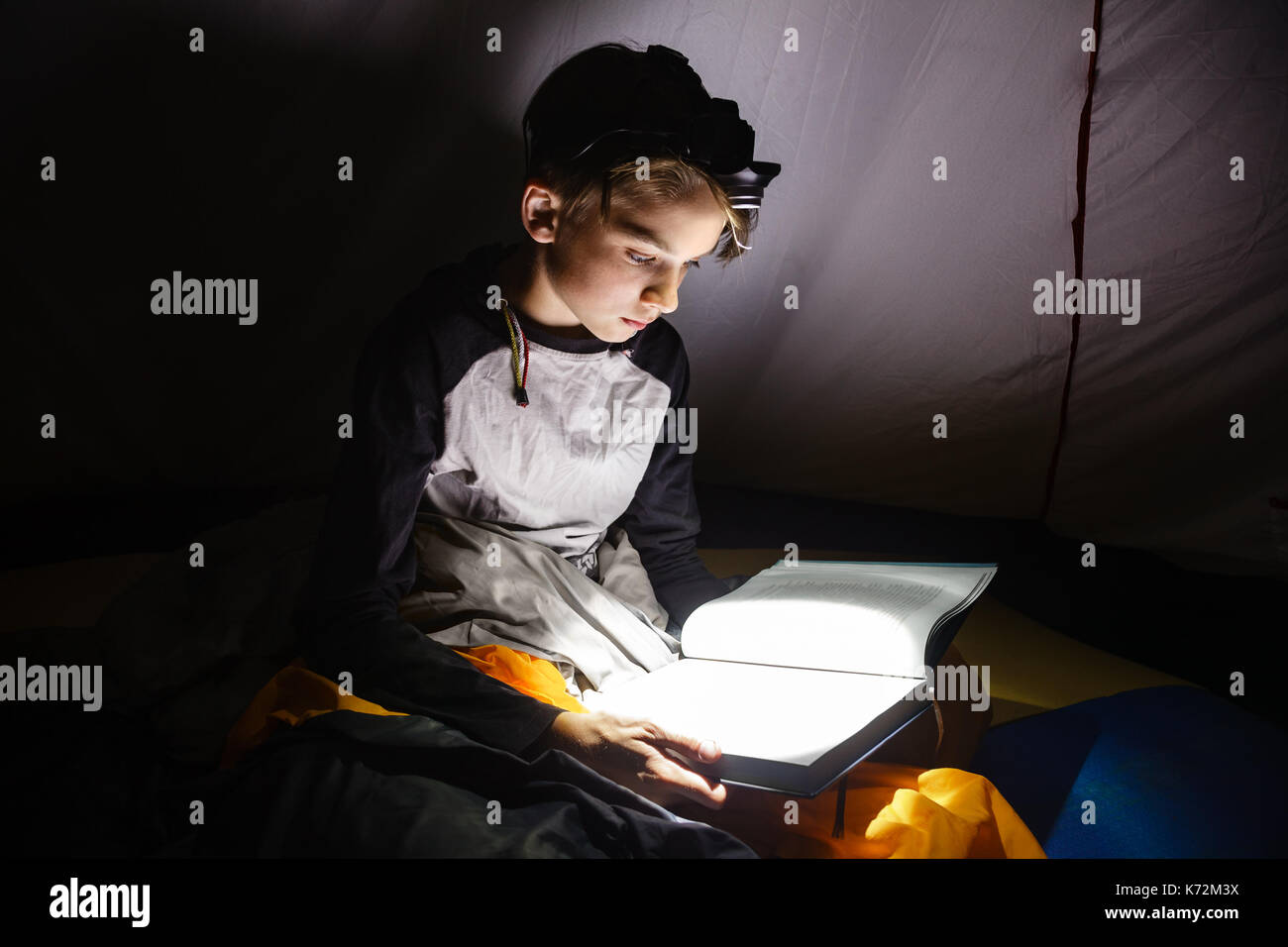 Teen boy sitting in a camping tent wrapped in a sleeping bag reading a
