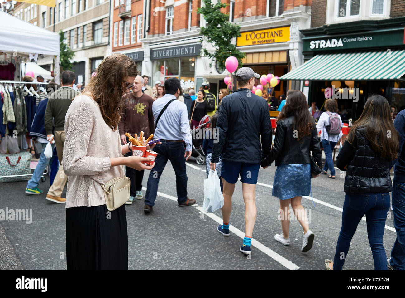Woman on mobile phone cellphone texting whilst eating churros food on a London street with people UK Stock Photo