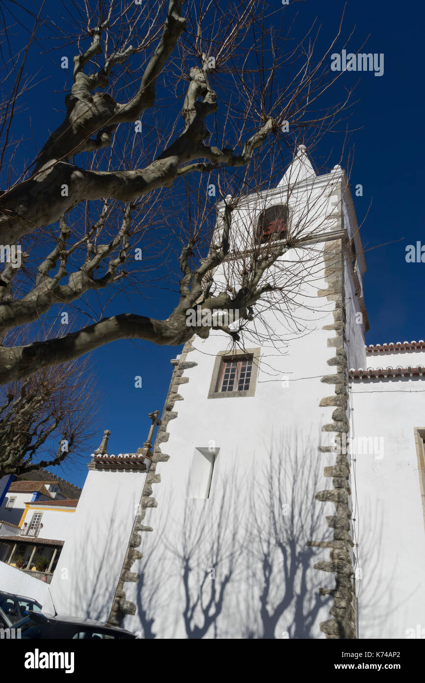 Low angle of the Church of Santa Maria framed by a gnarled leafless tree, in the medieval town of Obidos, Portugal, Stock Photo