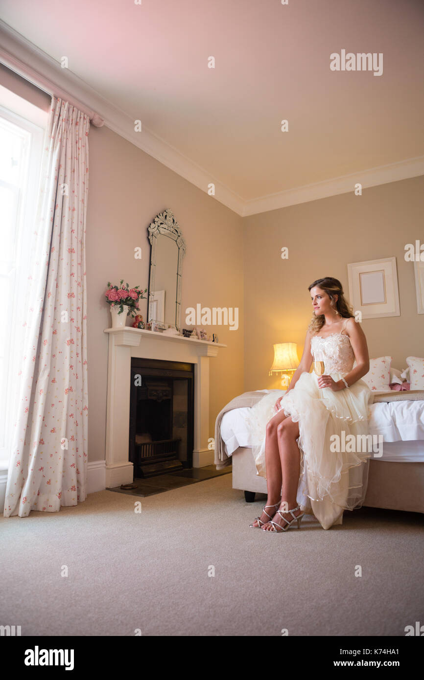 Full length of thoughtful bride holding champagne looking away while sitting on bed at home Stock Photo