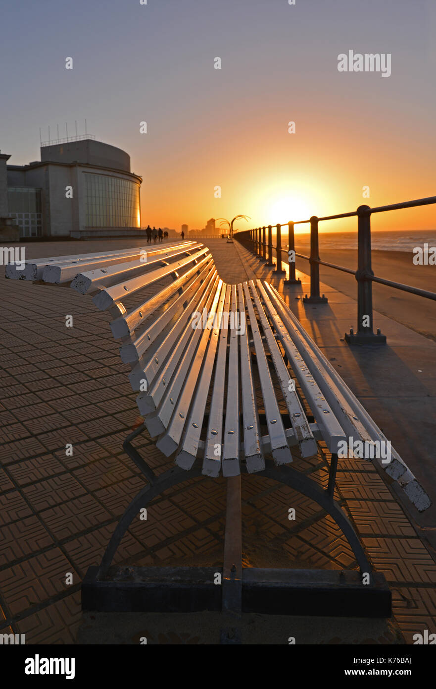Sunset along the waterfront promenade with a bench in the foreground in Ostend by its North Sea beach with a skyline view in the background, Belgium. Stock Photo