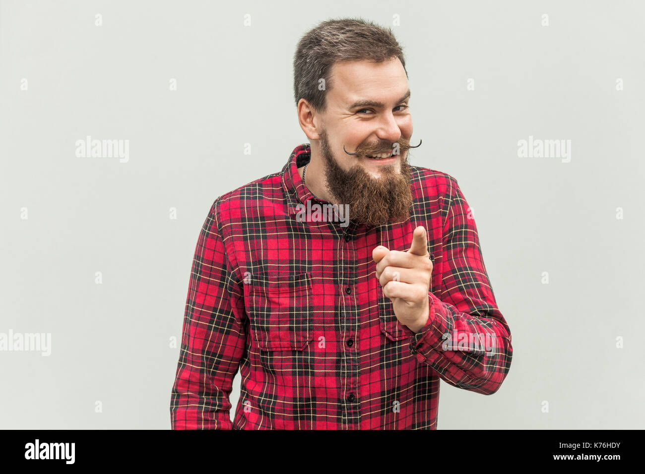 Shame of you! Bearded man in red shirt pointing finger at camera and toothy smile. Isolated on gray background, studio shot Stock Photo