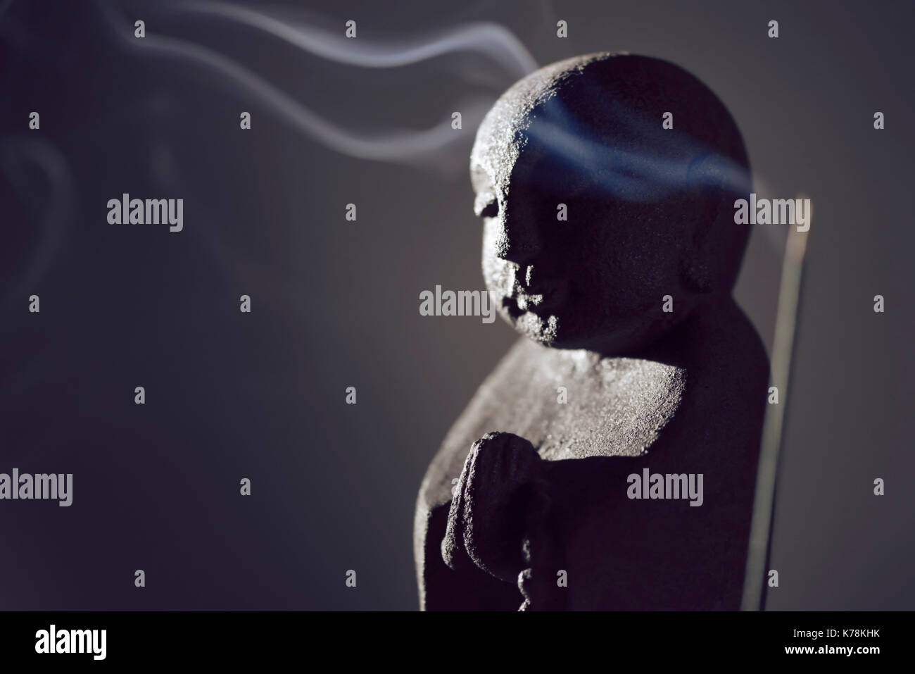 Praying Buddha statue with burning incense smoke in dramatic dim light on dark gray background Stock Photo